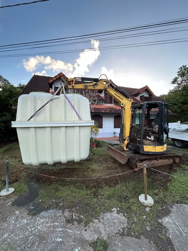 La cuve de micro-station posée sur la pelouse, la pelle jaune derrière, la maison au fond.